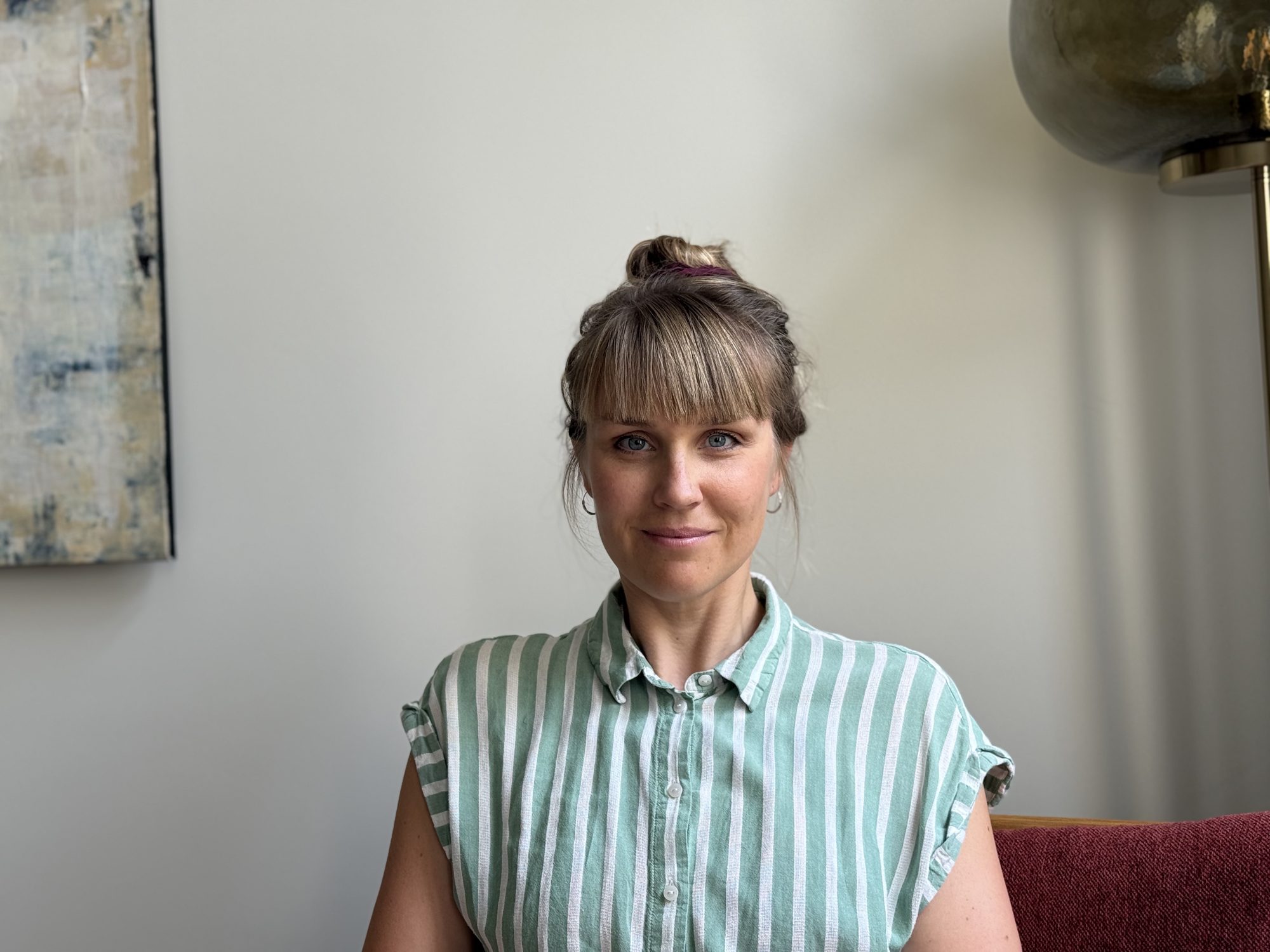 Alena Knabe-Lorenz, sitzend, vor einer hellen Wand mit abstraktem Kunstwerk und einer Stehlampe.